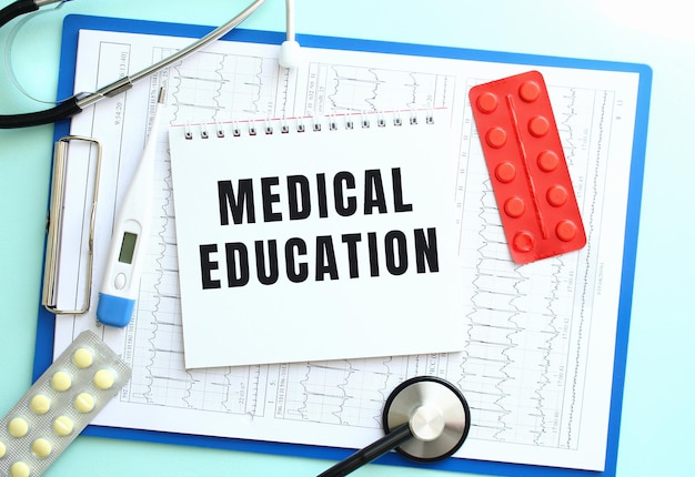 A notepad with the text medical education lies on a medical clipboard with a stethoscope and pills o