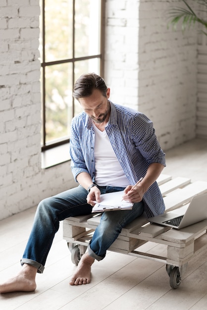 Noting down ideas. Nice serious handsome man sitting in the room and leaning over his notes while writing something with his pencil