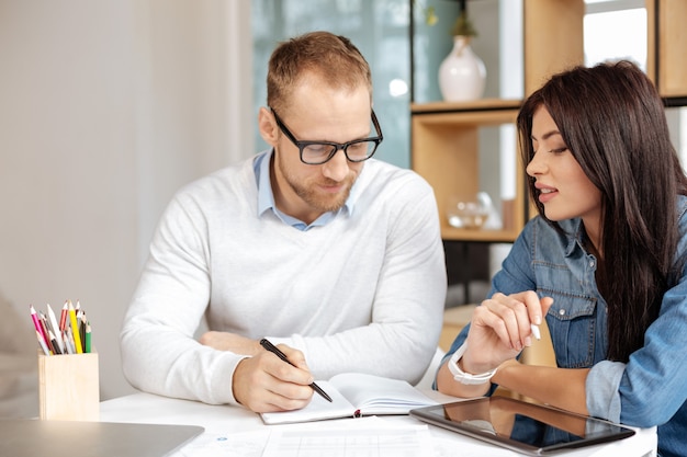 Noting down a plan. Intelligent handsome nice man sitting together with his colleague and looking at the notebook while developing a plan for future work