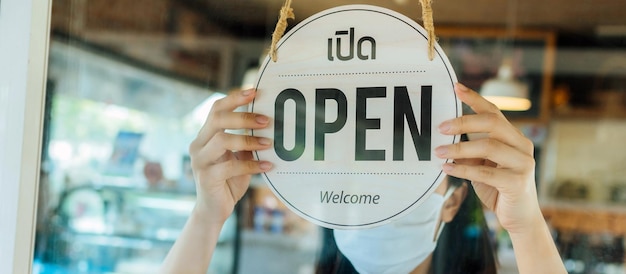 Now Open. asian woman staff wearing protection face mask turning Open sign board with Thai language on glass door in cafe coffee shop, cafe restaurant, small business owner, food and drink concept