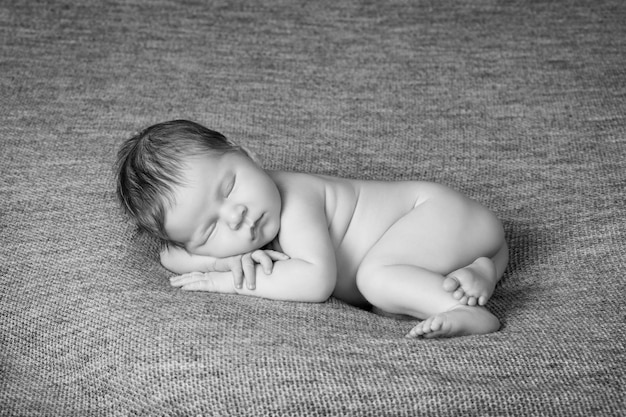 A nude newborn baby curled up and asleep on a gray textured blanket