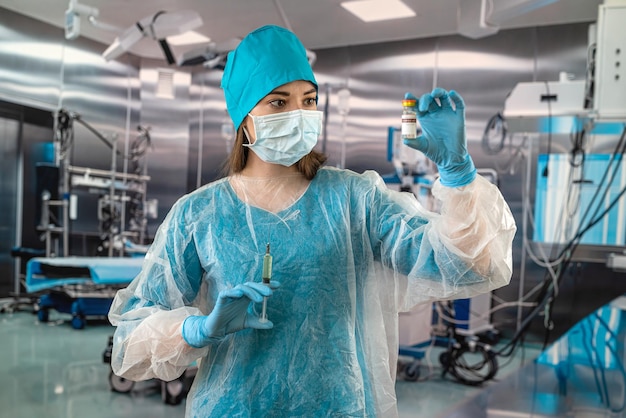 Nurse in a blue uniform with a mask and gloves behind a hat holding a syringe with medicine