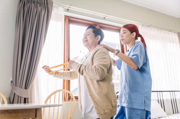 Nurse caregiver wearing scrubs exercises with a senior Asian woman by using resistance band exercise for the senior patient in physiotherapy treatment Home health care and nursing home concept