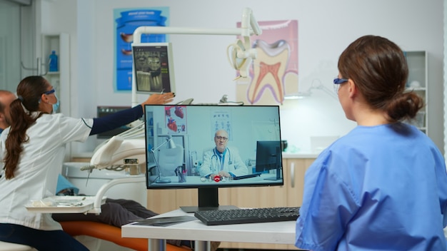 Nurse discussing about consultation with senior dentist medic on video call in dental office, while woman doctor is working with patient in background. Assistent listening medic on webcam