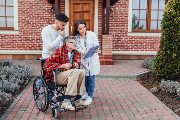 Nurse looking  on a folder ,old man in whelchair with his son waiting