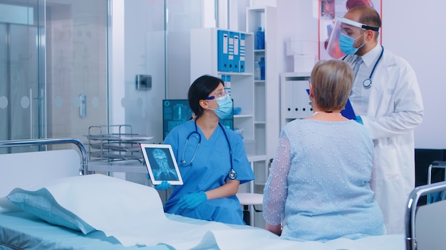 Nurse in mask explaining diagnostic on digital tablet PC to old retired senior woman while the doctor is entering hospital room to check on them. Healthcare system during or after coronavirus outbreak