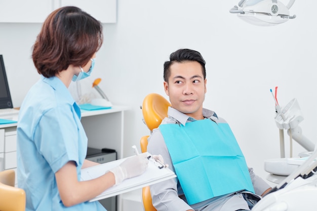 Nurse in medical mask talking to patient sitting in dental chair and telling his complaints