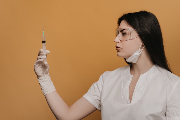 Nurse in protective glasses and a dressing scrubs, holding a syringe is about to inject a patient with coronavirus, over yellow background.