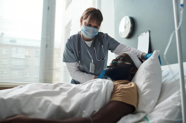 Nurse in protective mask straightening pillow of injured patient