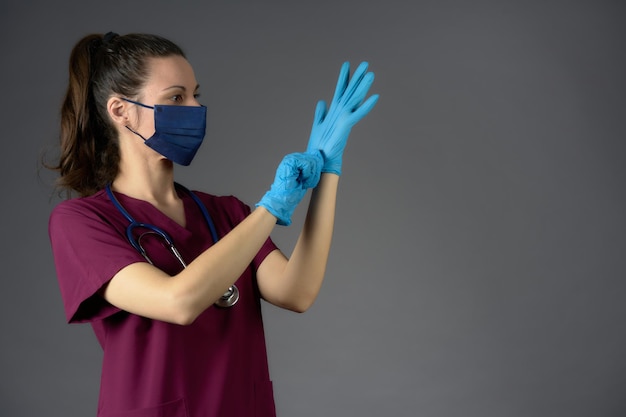 Nurse in purple uniform mask and stethoscope putting on latex medical gloves on gray background