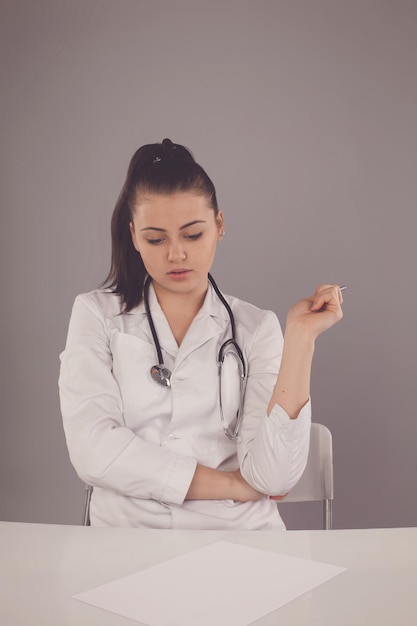 Nurse in white robe and with stethoscope on her neck is making a report at the table against