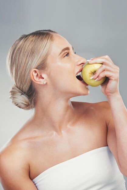 Nutrition smile and woman eating an apple for health of teeth dental and care for body against a grey studio background Food diet and happy model thinking of healthy lifestyle with a fruit