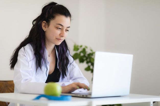 Nutritionist doctor sitting at her cabinet in weightloss clinic using laptop giving online consultation about healthy eating and meal plan program