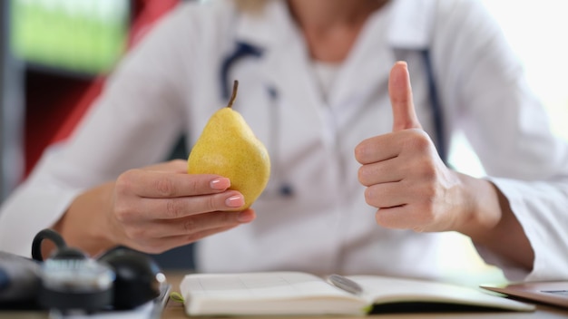 Nutritionist holding fresh pear fruit in hand and show thumbs up gesture female doctor gives