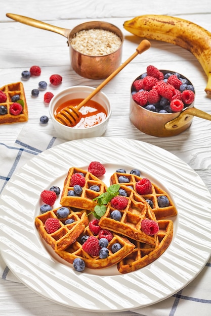 Oatmeal banana waffles served with berries and honey on a white plate on a wooden table with ingredients, berries and honey, vertical view from above,