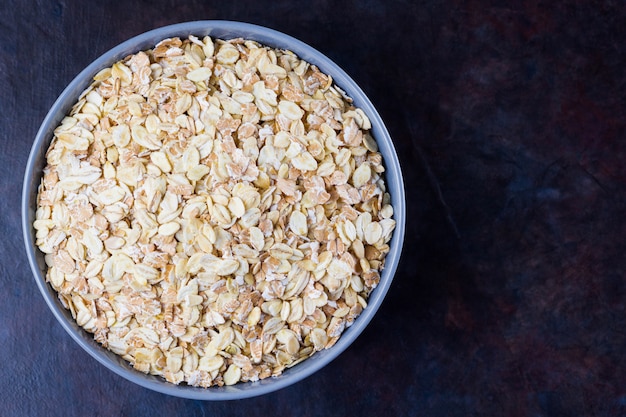 Oatmeal in bowl. Oat flakes on dark background. Cereals for making healthy breakfast. Vegetarian food