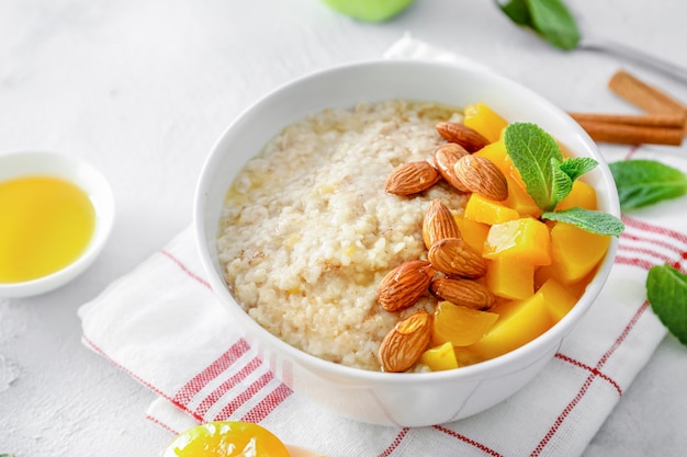 Oatmeal bowl with mint leaves, cinnamon, peach pieces, almond nuts and honey on white background.