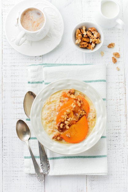 Oatmeal porridge with caramelized persimmon and walnuts in a light ceramic bowl on a white wooden table. Selective focus. Top view. Copy space.