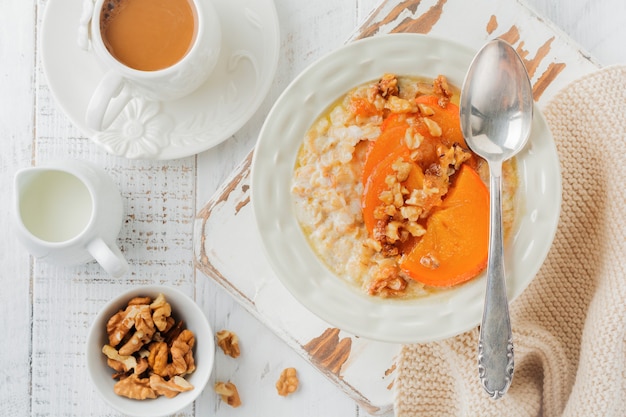 Oatmeal porridge with caramelized persimmon and walnuts in a light ceramic bowl on a white wooden table. Selective focus. Top view. Copy space.