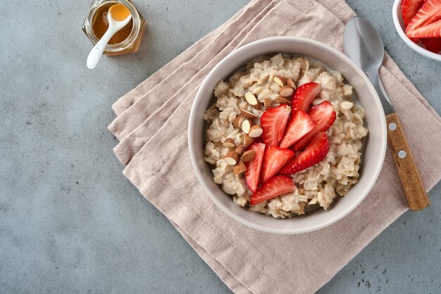 Oatmeal porridge with strawberry slices, nuts almonds and honey in bowl on grey table. Healthy eating, dieting, vegetarian food concept. Place for text.