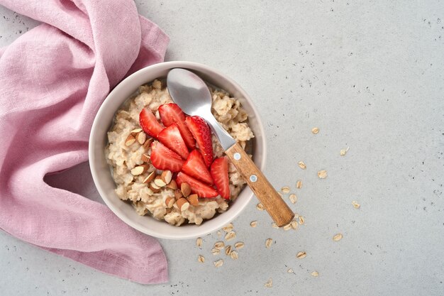 Oatmeal porridge with strawberry slices, nuts almonds and honey in bowl on grey table. Healthy eating, dieting, vegetarian food concept. Place for text.