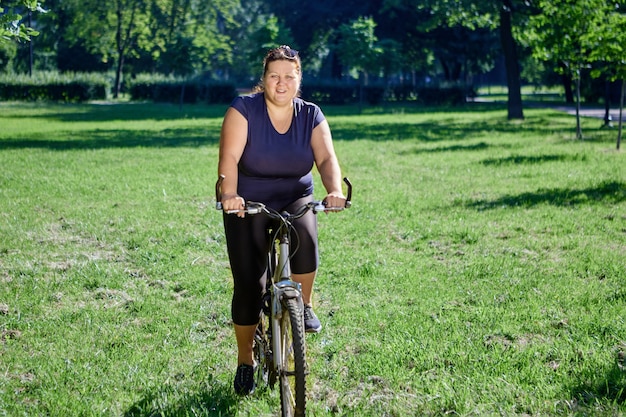 Obese woman in body positivity style rides bicycle on lawn on sunny summer day