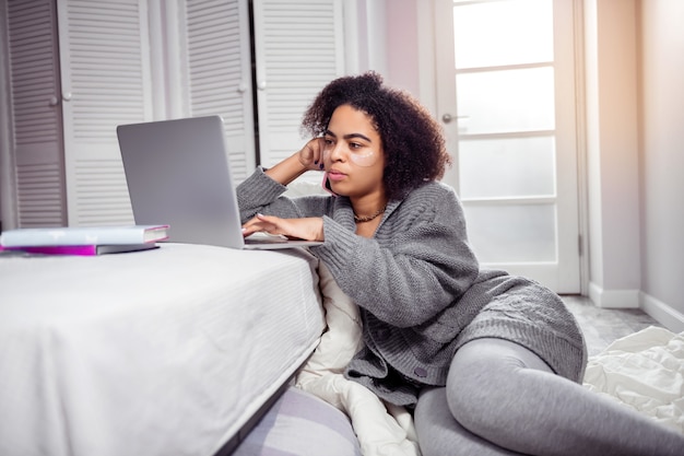 Occupied with laptop. Attentive curly woman wearing cozy cloth and tying on laptop keyboard while having workday at home