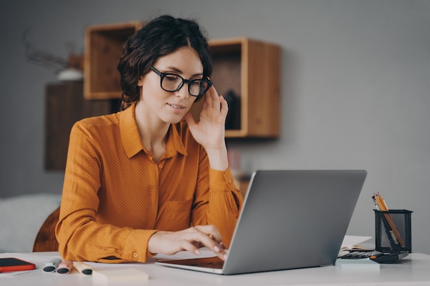 Ocused spanish woman in glasses sits at desk and typing on laptop during remote work at home