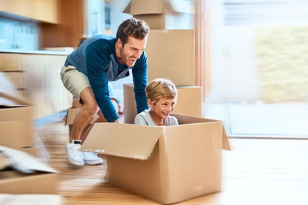 Off we go in the spaceship Shot of a cheerful young man pushing his son around in a box imagining its a car inside at home during the day