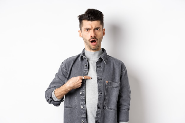 Offended young man being accused, frowning and pointing at himself shocked, standing on white background.