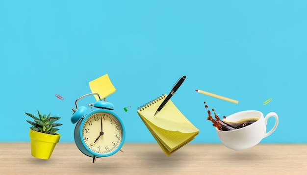 Office accessories notepad, alarm clock, coffee cup, plant, pen flying over desk table on blue wall background. Mockup with copy space