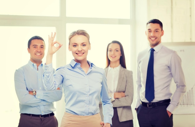 office, business and teamwork concept - friendly young smiling businesswoman with team on back showing ok-sign