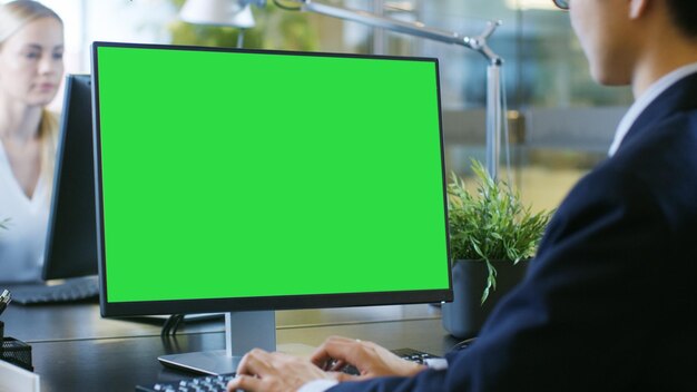 Photo in the office businessman works at his desk on a personal computer with mockup green screen his colleague sets opposite to him