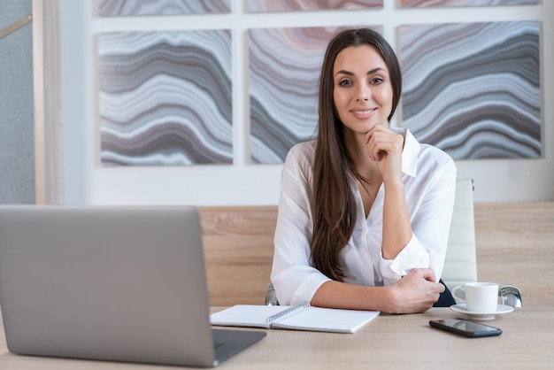 Office manager woman wearing white shirt smiling sitting to table with laptop and cup of coffee looking at the camera Happy office worker in big light office room Concept of work