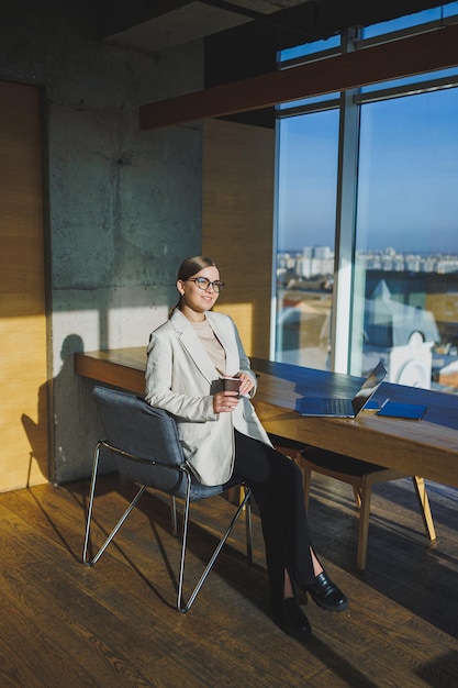 Office work attractive young female manager working in office standing in bright workspace using modern laptop computer young woman drinking delicious coffee