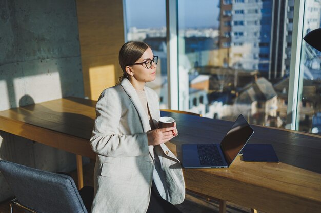 Office work attractive young female manager working in office standing in bright workspace using modern laptop computer young woman drinking delicious coffee