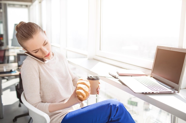 Office worker has some lunch. Girl has a croissan and a cup of coffee to eat ad drink. Also woman is talking on the phone. She put the phone between her head and shoulder because her hands are busy