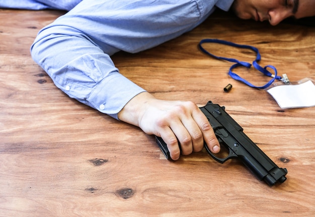 Office worker lies on the floor holding gun in his hands. Suicide because of work stress. Depression or burnout. Terrible life situation. Man near the laptop at the desk.