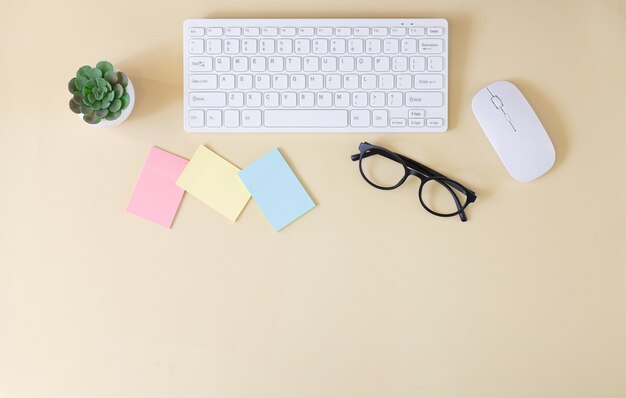 Office workplace table with computer keyboard, mouse, glasses, blank stickers and plant top view