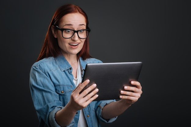 Oh my gosh. Cute fun adorable lady expressing her feelings while reading something on her tablet and standing isolated on grey background