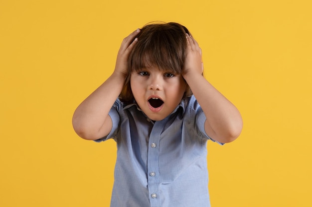 Oh no Cute little boy having some troubles touching his head with shock looking at camera with open mouth