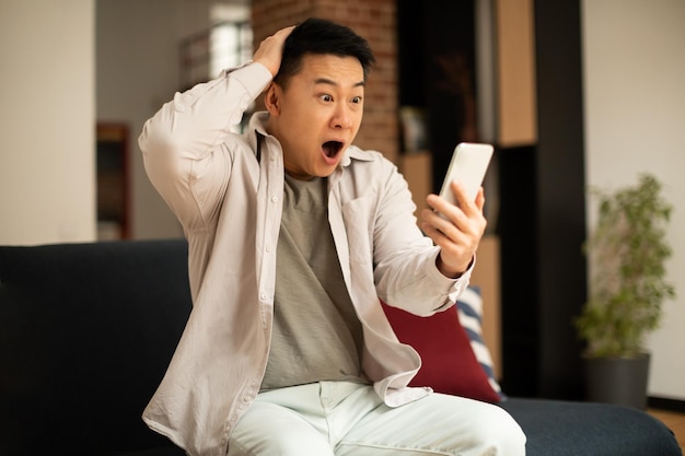 Oh no Shocked mature asian man reading bad message on smartphone while sitting on sofa at home free space