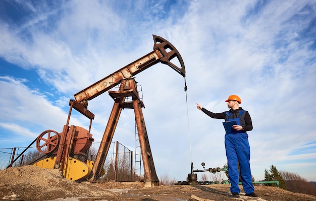 Oil worker in uniform and helmet working in an oilfield next to a pump jack on a sunny day