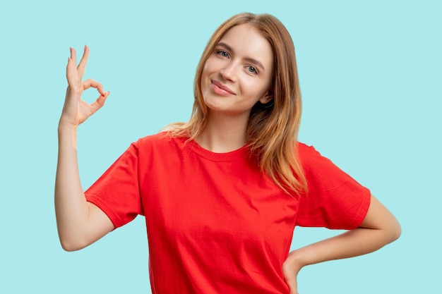 OK sign Well done Portrait of satisfied friendly woman in red tshirt encouraging with approval hand gesture smiling isolated on blue background Deal agreement Support acceptance