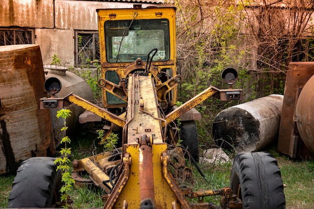 Old abandoned broken road construction machine (yellow motor grader). Rusty metal, broken glass, missing headlights and remnants of old yellow paint