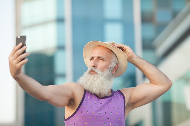 Old active bearded man in hat taking selfie with mobile phone on street.