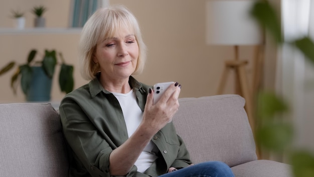 Old Caucasian woman 60s elderly mature lady sits at home on sofa with mobile phone reads news