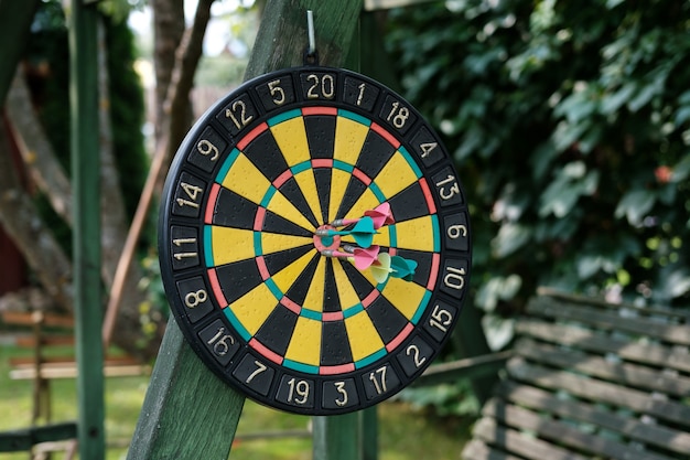 An old circular dart board with plastic darts hangs on a wooden structure in the garden.