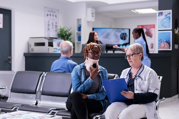 Old doctor consulting woman at facility, wearing cervical neck collar to cure injury and pain. Specialist and patient with medical foam brace talking about recovery in waiting room at health center.