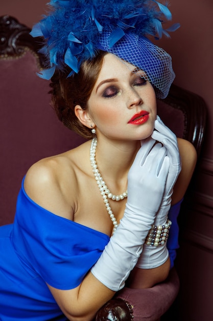 Old Fashioned retro great britain style photography. Beautiful young caucasian model in blue dress and fashion makeup and hat posing sitting on the artificial sofa and looking into the distance.